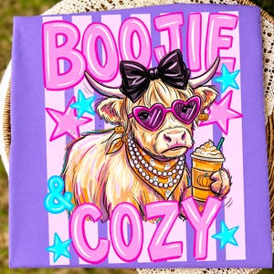 May include: A purple t-shirt featuring a graphic of a highland cow wearing a black bow, heart-shaped sunglasses, and a ghost-patterned bandana. The cow holds a coffee cup, surrounded by pink stars and the text BOOJIE & COZY in pink letters.