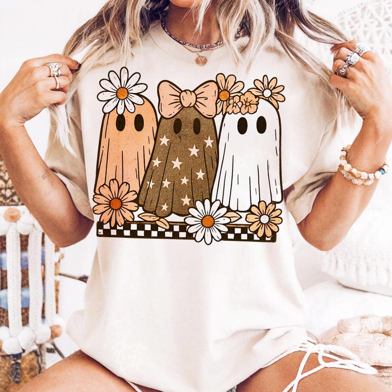 May include: A beige t-shirt featuring a graphic of three cartoon ghosts with daisy accents. The ghosts are in shades of orange, brown, and white, with a bow and star details. The design is set against a black and white checkered border.