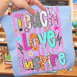 Puede incluir: Una sudadera azul claro con un estampado gr&aacute;fico con las palabras Teach, Love, Inspire. El dise&ntilde;o incluye rayas a cuadros rosas, patrones coloridos, flores, estrellas, rayos y motivos de l&aacute;pices en tonos rosa, naranja y azul.
