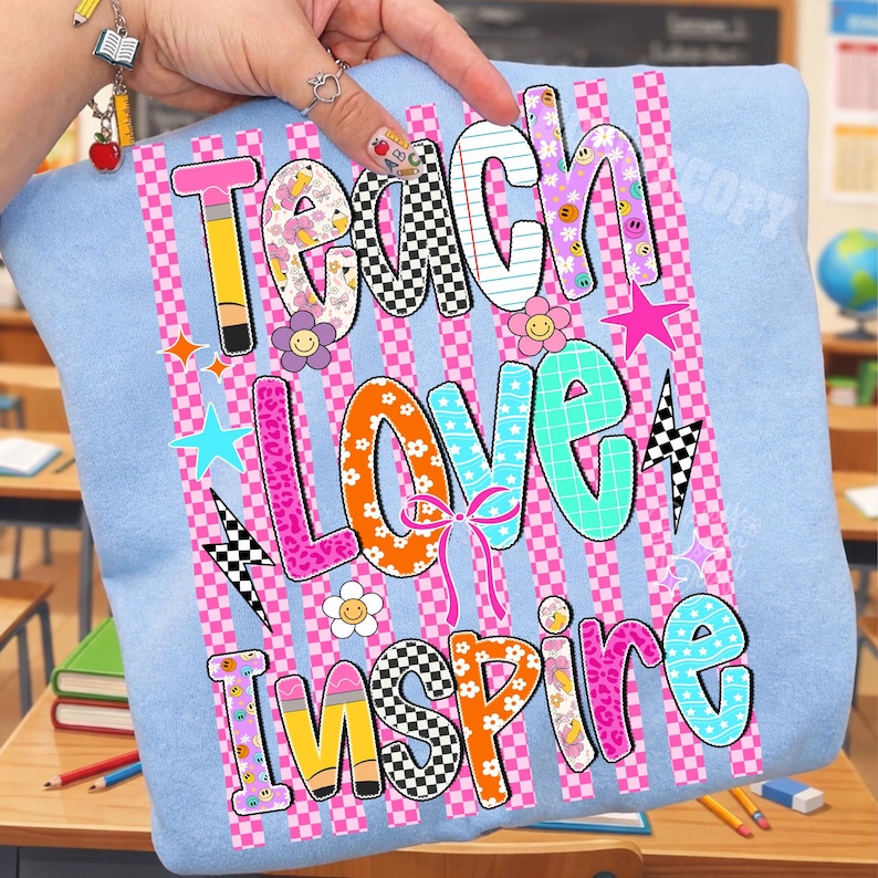 Puede incluir: Una sudadera azul claro con un estampado gr&aacute;fico con las palabras Teach, Love, Inspire. El dise&ntilde;o incluye rayas a cuadros rosas, patrones coloridos, flores, estrellas, rayos y motivos de l&aacute;pices en tonos rosa, naranja y azul.
