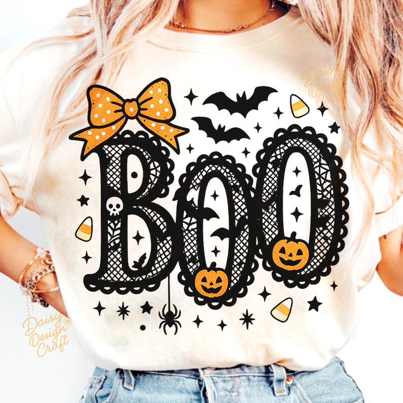 Puede incluir: Camiseta color crema con un dise&ntilde;o tem&aacute;tico de Halloween. La palabra "BOO" se muestra en letras negras ornamentadas, adornada con un lazo, murci&eacute;lagos, calabazas, estrellas, caramelos y una ara&ntilde;a. Para la temporada de Halloween.