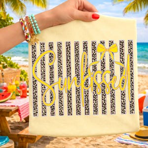 以下が含まれることがあります： 黒とヒョウ柄のストライプと、黄色のスクリプトで「SunKissed」という文字が入った、明るい黄色のビーチタオル。 黄色のリボンが単語の上にあります。 タオルは、手首にブレスレットをつけた人が持っています。