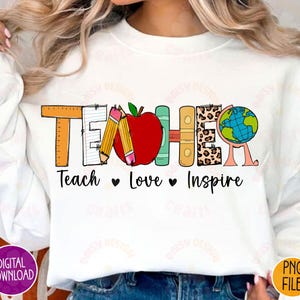 K&ouml;nnte beinhalten: Wei&szlig;er Pullover mit einem Grafikdesign, das "Teacher" in bunten Buchstaben sagt. Die Buchstaben bestehen aus verschiedenen Schulmaterialien, darunter Bleistifte, ein Lineal, ein Buch und ein Globus. Der Text "Teach Love Inspire" befindet sich unter der Grafik.