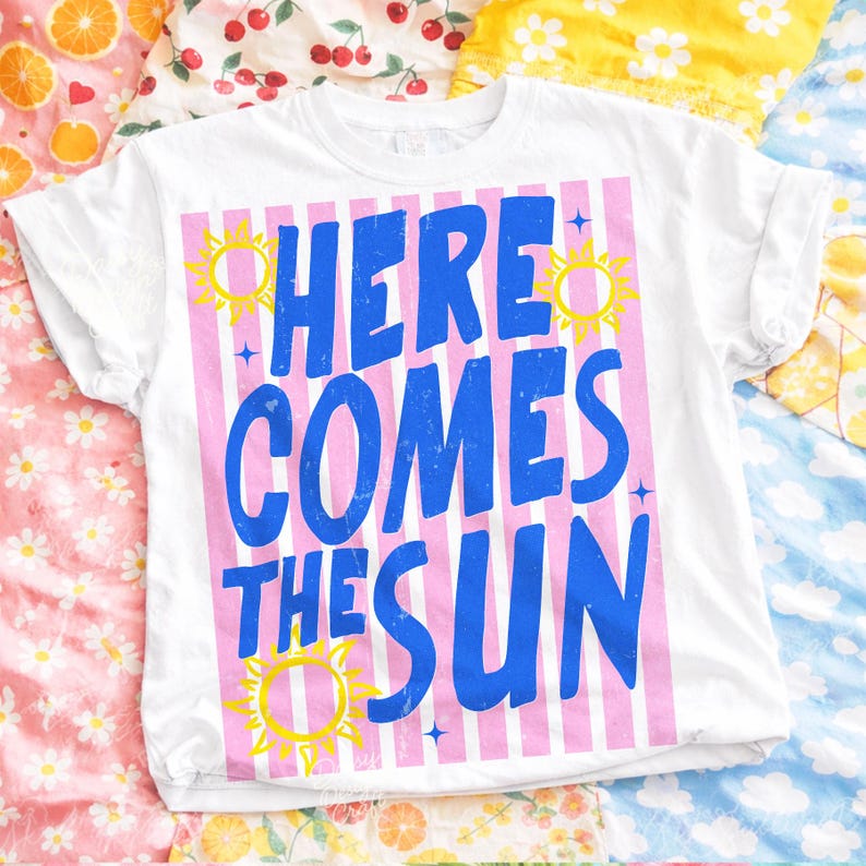 May include: White t-shirt with rolled sleeves featuring the text "HERE COMES THE SUN" in blue, set against pink vertical stripes. Yellow sun illustrations and blue stars accent the design. The shirt is displayed on a colorful patterned surface.