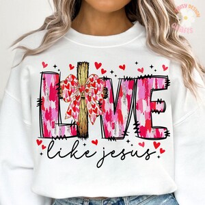 May include: White sweatshirt with the word "LOVE" in pink brushstroke letters, a wooden cross with a bow, and the phrase "like jesus" in cursive. Red hearts and black stars decorate the design.