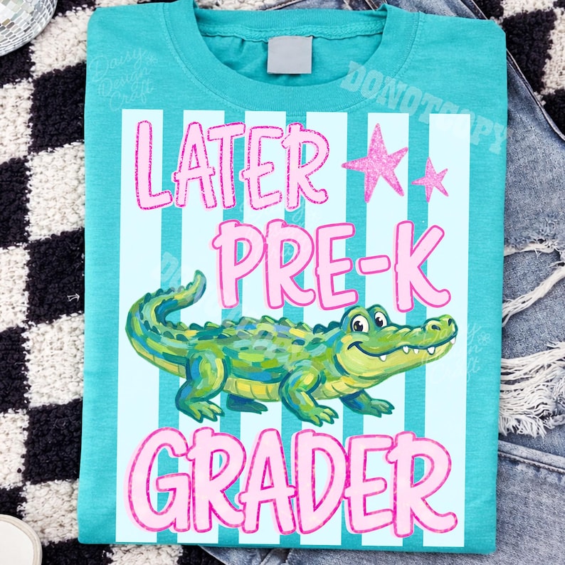 以下が含まれることがあります： 水色と白のストライプ模様の、ティール色のTシャツ。「LATER PRE-K GRADER」のピンク色の文字と、漫画のアリゲーターのイラスト、2つのピンク色の星が特徴です。
