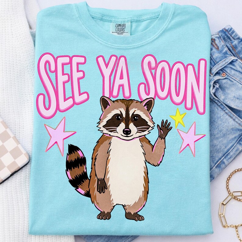 K&ouml;nnte beinhalten: Hellblaues T-Shirt mit dem Schriftzug "SEE YA SOON" in Rosa. Ein Cartoon-Waschb&auml;r winkt, begleitet von rosa und gelben Sternen. Das T-Shirt ist aus weichem Material.
