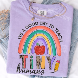 Puede incluir: Una camiseta de color morado claro con un gr&aacute;fico de un arco&iacute;ris colorido, una manzana roja y un l&aacute;piz amarillo. El texto dice "ITS A GOOD DAY TO TEACH TINY HUMANS". La palabra "TINY" est&aacute; escrita con un estampado de leopardo. Tiene cuello redondo.