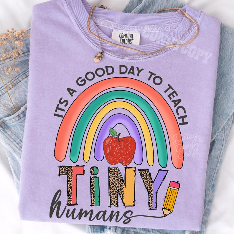 Puede incluir: Una camiseta de color morado claro con un gr&aacute;fico de un arco&iacute;ris colorido, una manzana roja y un l&aacute;piz amarillo. El texto dice "ITS A GOOD DAY TO TEACH TINY HUMANS". La palabra "TINY" est&aacute; escrita con un estampado de leopardo. Tiene cuello redondo.