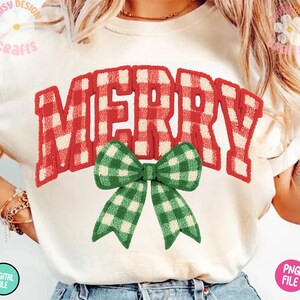 May include: Cream-colored t-shirt with the word "MERRY" in red and white plaid lettering. A green and white plaid bow is centered below the word. The shirt has a casual, festive design, perfect for the holiday season.