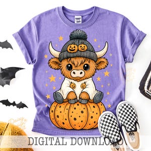 Op de afbeelding: Een paars T-shirt met een cartoon Highland-koe met een pompoenhoed en zittend op een pompoen. Het ontwerp bevat sterren en de tekst "DIGITAL DOWNLOAD". Een paar zwart-wit geruite instappers liggen ernaast.
