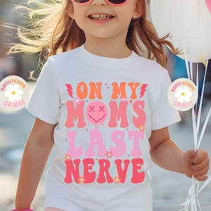 Op de afbeelding: Wit T-shirt met roze en oranje tekst die "On My Mom's Last Nerve" luidt. De tekst is in een retro lettertype stijl met een druppelend smiley gezicht.