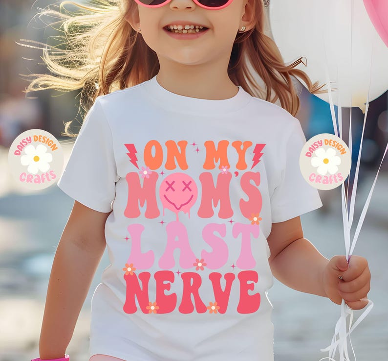 Op de afbeelding: Wit T-shirt met roze en oranje tekst die "On My Mom's Last Nerve" luidt. De tekst is in een retro lettertype stijl met een druppelend smiley gezicht.