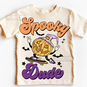 May include: A cream-coloured t-shirt with a Halloween graphic. The design shows a smiling jack-o'-lantern wearing a witch's hat and skateboarding. The words "Spooky Dude" are written in orange and purple.