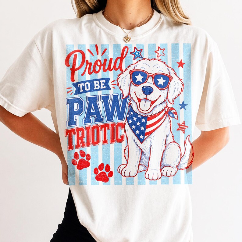 May include: Cream-colored t-shirt with a patriotic graphic. The design features a smiling white dog wearing star-shaped sunglasses and a bandana with a US flag pattern. The text reads "Proud to be PAW TRIOTIC" in red and blue.