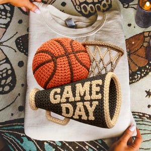 K&ouml;nnte beinhalten: Beiges Sweatshirt mit einem H&auml;keldesign eines Basketballs, Netzes und Megaphons. Auf dem Megaphon steht "GAME DAY" in cremefarbenen Buchstaben. Der Basketball ist orange und braun, das Netz beige.