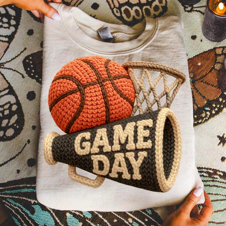 K&ouml;nnte beinhalten: Beiges Sweatshirt mit einem H&auml;keldesign eines Basketballs, Netzes und Megaphons. Auf dem Megaphon steht "GAME DAY" in cremefarbenen Buchstaben. Der Basketball ist orange und braun, das Netz beige.