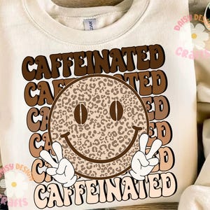 Puede incluir: Una sudadera blanca con un dise&ntilde;o gr&aacute;fico marr&oacute;n y beige que presenta una cara sonriente con granos de caf&eacute; como ojos y el texto "CAFFEINATED" repetido en una fuente retro.