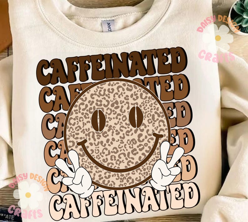 Puede incluir: Una sudadera blanca con un dise&ntilde;o gr&aacute;fico marr&oacute;n y beige que presenta una cara sonriente con granos de caf&eacute; como ojos y el texto "CAFFEINATED" repetido en una fuente retro.