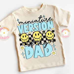 Op de afbeelding: Een beige T-shirt met de tekst "miniature version of my dad" in blauw en geel. De tekst wordt omringd door smiley-gezichten en een geruit patroon.