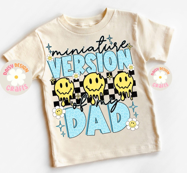 Op de afbeelding: Een beige T-shirt met de tekst "miniature version of my dad" in blauw en geel. De tekst wordt omringd door smiley-gezichten en een geruit patroon.