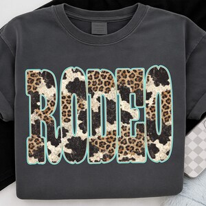 Pu&ograve; includere: T-shirt grigio scuro con la scritta "RODEO" in grandi lettere consumate. Le lettere presentano un mix di motivi leopardati e muccati con un contorno azzurro. La maglietta ha maniche corte e girocollo.