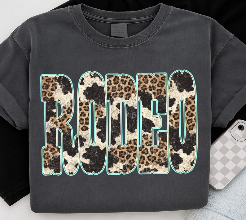 Pu&ograve; includere: T-shirt grigio scuro con la scritta "RODEO" in grandi lettere consumate. Le lettere presentano un mix di motivi leopardati e muccati con un contorno azzurro. La maglietta ha maniche corte e girocollo.