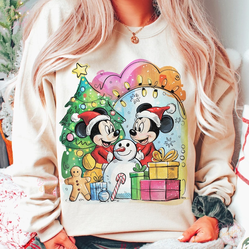May include: A beige sweatshirt featuring a festive Christmas design. The graphic includes Mickey and Minnie Mouse in Santa hats, a snowman, a Christmas tree, and wrapped gifts. The design is colorful with a cartoon style.