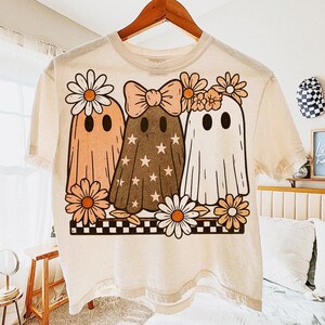 May include: Cream-colored t-shirt featuring a graphic of three cartoon ghosts with daisy accents. The ghosts are in shades of peach, brown, and white. One ghost has a bow, and another has a daisy crown. The design is set on a black and white checkered border.