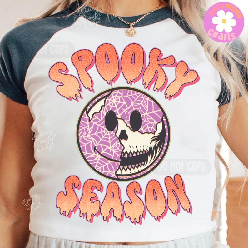 May include: White raglan t-shirt with navy sleeves. The shirt displays the words "Spooky Season" in orange and pink dripping letters. A purple smiley face with a spiderweb design and a skull graphic is in the centre.