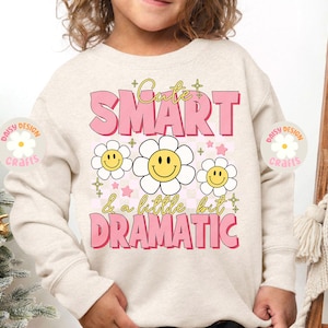 Op de afbeelding: Een licht beige sweatshirt met de tekst "Cute Smart & a little bit Dramatic" in roze letters. De tekst wordt omringd door gele smiley-gezichten met witte madeliefjes.