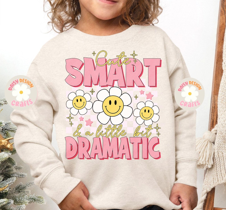 Op de afbeelding: Een licht beige sweatshirt met de tekst "Cute Smart & a little bit Dramatic" in roze letters. De tekst wordt omringd door gele smiley-gezichten met witte madeliefjes.