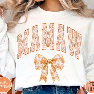 May include: White sweatshirt with the word "MAMAW" in orange letters with a floral pattern. The letters are outlined in black. Below the word is a large orange bow with a floral pattern.