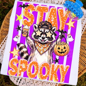 May include: A white t-shirt featuring a graphic of a raccoon wearing a baseball cap, star-shaped sunglasses, and a ghost-patterned bandana. The raccoon holds a coffee cup and a jack-o'-lantern bucket. The text reads STAY SPOOKY in orange.