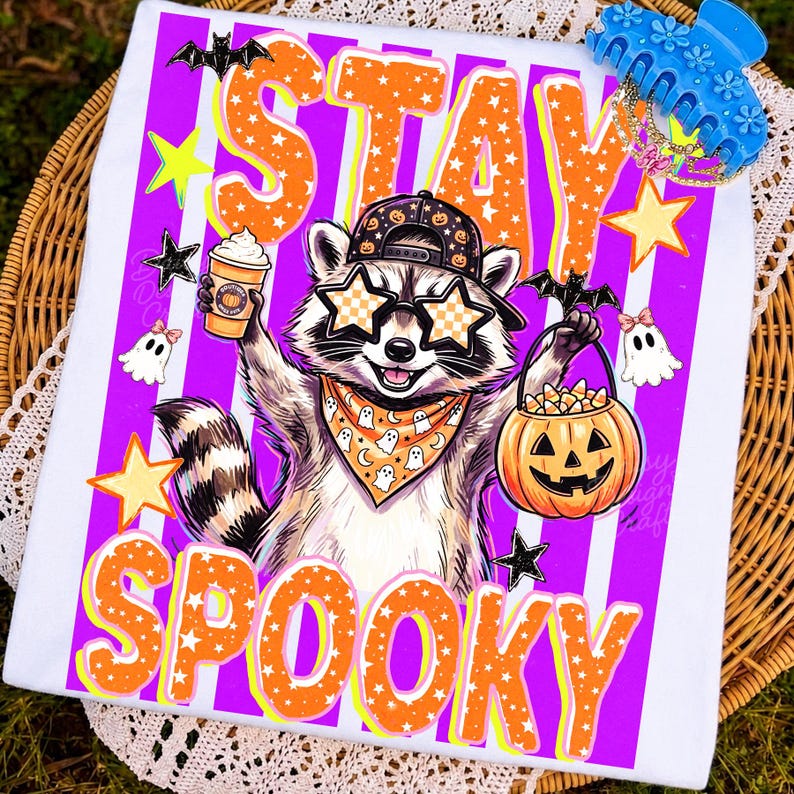 May include: A white t-shirt featuring a graphic of a raccoon wearing a baseball cap, star-shaped sunglasses, and a ghost-patterned bandana. The raccoon holds a coffee cup and a jack-o'-lantern bucket. The text reads STAY SPOOKY in orange.