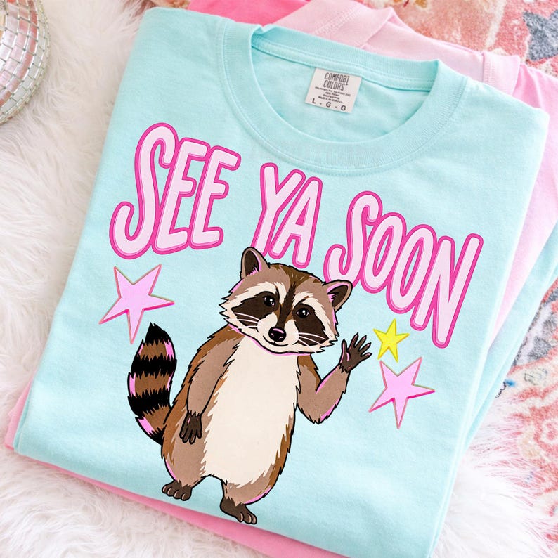 K&ouml;nnte beinhalten: Hellblaues T-Shirt mit dem Schriftzug "SEE YA SOON" in Rosa und einem winkenden Cartoon-Waschb&auml;ren. Rosa und gelbe Sterne akzentuieren das Design. Das T-Shirt liegt gefaltet auf einem rosa T-Shirt.