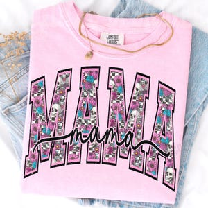 May include: Pink Comfort Colors sweatshirt with the word "MAMA" in a black, block letter design. The letters are filled with a checkerboard pattern, skulls, and floral accents. A gold necklace rests on the shirt.