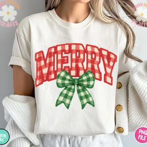 May include: White t-shirt featuring the word "MERRY" in red and white gingham print, with a green and white gingham bow. The shirt is a casual style, perfect for the holiday season.