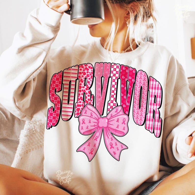 May include: A cream-colored sweatshirt with the word "SURVIVOR" in pink, featuring various patterns and a pink ribbon bow. The design includes hearts, ribbons, and checkered patterns. The sweatshirt is a casual garment.