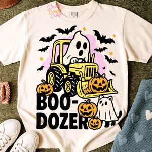 Op de afbeelding: Cr&egrave;mekleurig T-shirt met een Halloween-thema. Het ontwerp bevat een gele tractor met een spookchauffeur, pompoenen, vleermuizen en de woorden "BOO-DOZER". Een spookkat is ook aanwezig.