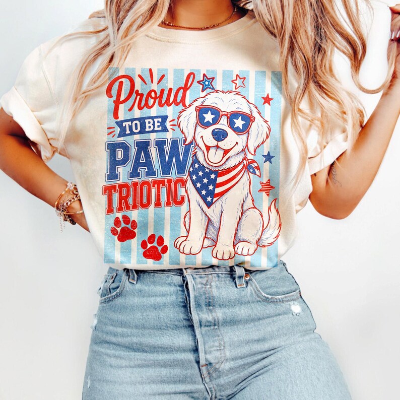 May include: Cream-colored t-shirt featuring a patriotic dog graphic. The dog wears sunglasses and a bandana with an American flag design. The shirt has the words "Proud to be PAWtriotic" in red and blue.