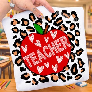 Puede incluir: Una camiseta blanca con un gr&aacute;fico de una manzana roja grande centrado sobre un fondo de estampado de leopardo negro y beige. La manzana est&aacute; decorada con contornos de corazones blancos y la palabra TEACHER en letras blancas.