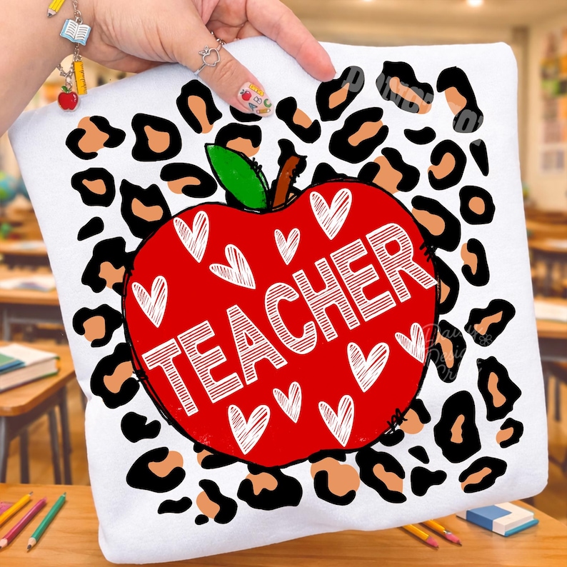Puede incluir: Una camiseta blanca con un gr&aacute;fico de una manzana roja grande centrado sobre un fondo de estampado de leopardo negro y beige. La manzana est&aacute; decorada con contornos de corazones blancos y la palabra TEACHER en letras blancas.
