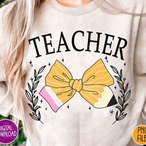 K&ouml;nnte beinhalten: Ein hellbeiger Pullover mit dem Wort "TEACHER" in Schwarz bedruckt. Eine gelbe Bleistiftschleife mit rosa und wei&szlig;en Akzenten ist mittig auf dem Pullover platziert. Die Schleife ist von schwarzen Blumenranken umgeben.