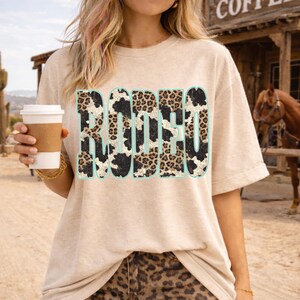 Pu&ograve; includere: T-shirt beige con la scritta "RODEO" a caratteri grandi e in grassetto. Le lettere presentano un design a stampa di pelle di mucca e leopardo con un contorno azzurro chiaro. La persona tiene in mano una tazza di caff&egrave;. Lo sfondo include un cavallo e un edificio.