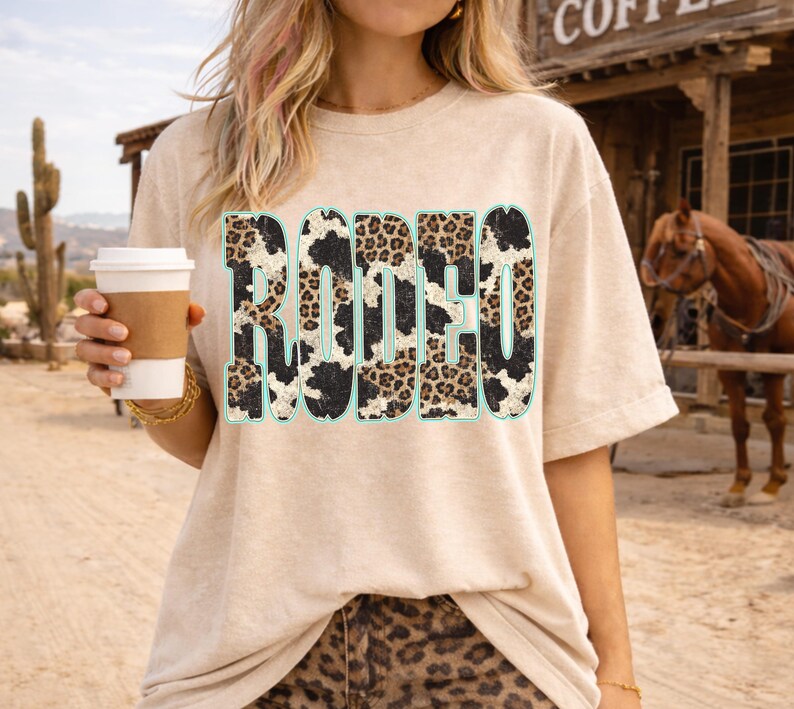 Pu&ograve; includere: T-shirt beige con la scritta "RODEO" a caratteri grandi e in grassetto. Le lettere presentano un design a stampa di pelle di mucca e leopardo con un contorno azzurro chiaro. La persona tiene in mano una tazza di caff&egrave;. Lo sfondo include un cavallo e un edificio.