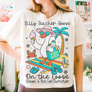 Puede incluir: Una camiseta de color crema con un gr&aacute;fico de un ganso con sombrero rosa y gafas de sol en forma de coraz&oacute;n. El ganso sostiene un cono de helado y una bolsa azul con la etiqueta Books & Beach. El texto dice Silly Teacher Goose On the Loose School's Out For Summer.