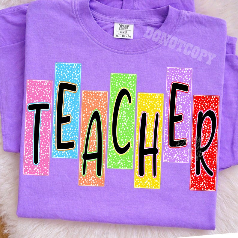 Puede incluir: Una camiseta de color morado claro con la palabra TEACHER en letras negras negritas. Cada letra est&aacute; colocada sobre un bloque rectangular vertical con un patr&oacute;n de motas blancas en colores rosa, azul, naranja, verde, amarillo, morado y rojo.