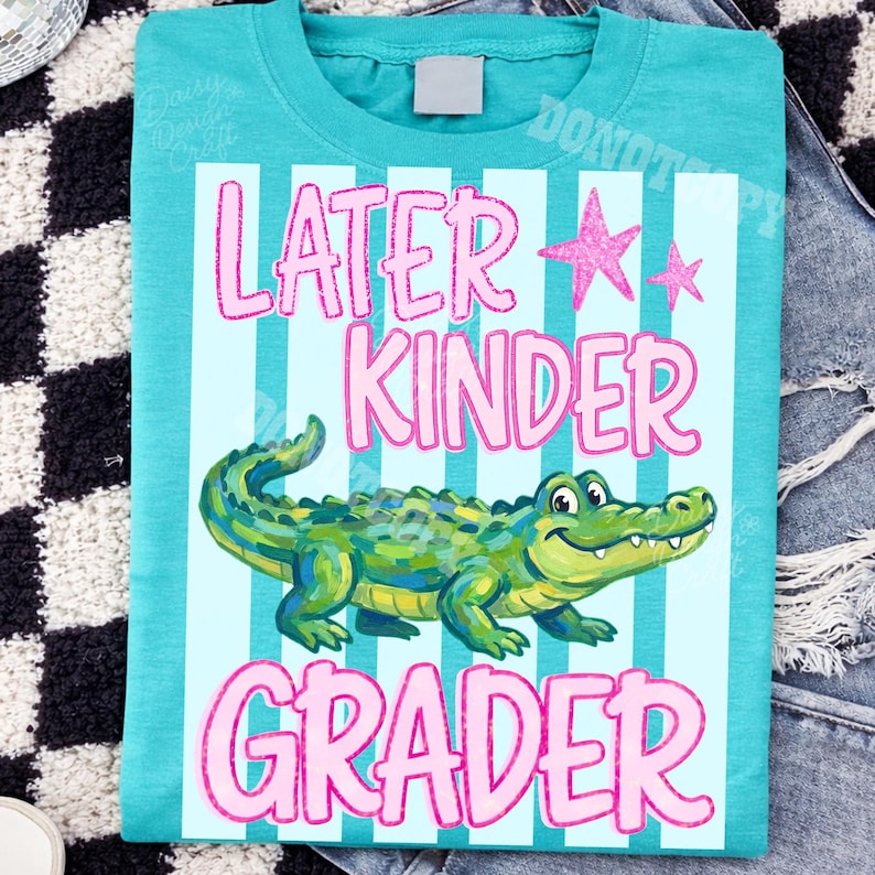 以下が含まれることがあります： ピンク色の「Later Kinder Grader」の文字と、漫画のアリゲーターのイラストが描かれた、ターコイズ色のTシャツ。デザインには、水色の縦縞とピンクの星のアクセントが含まれています。