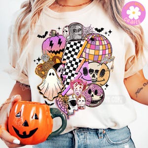 May include: Cream t-shirt with a Halloween graphic featuring a purple pumpkin, a checkered lightning bolt, a disco ball, skulls, and a ghost. An orange pumpkin mug is held in the foreground. The design is vibrant and festive.